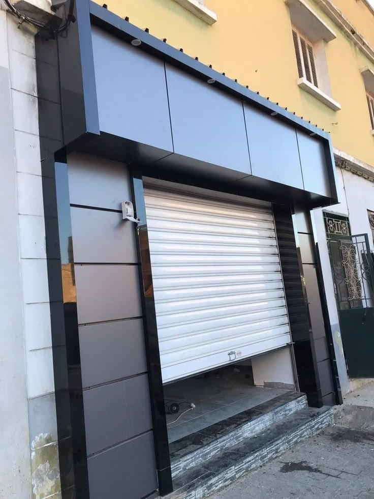 Commercial storefront with a silver metal roll-up door partially open, framed by modern black cladding and a raised entry threshold.