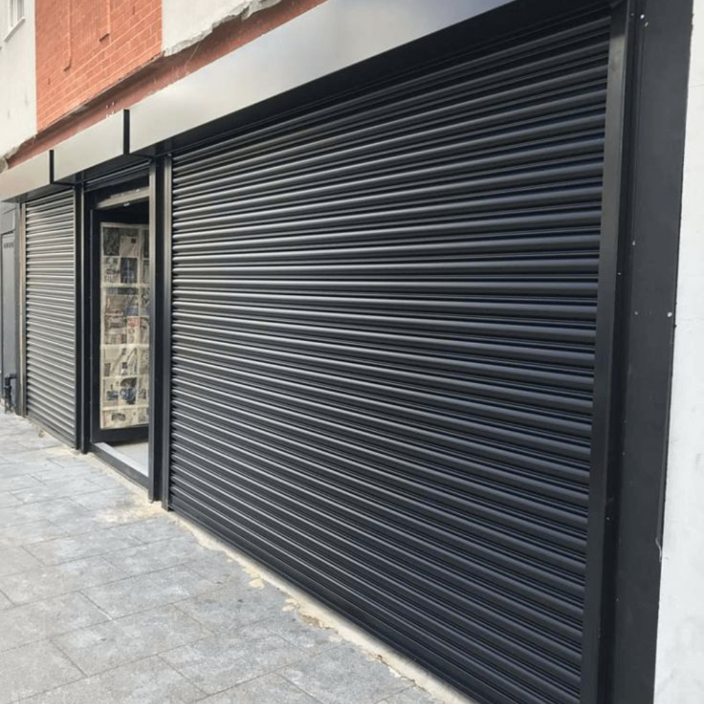 Commercial storefront with a black metal roll-up door installed beside a glass entry door, featuring ribbed shutter panels and a paved sidewalk.