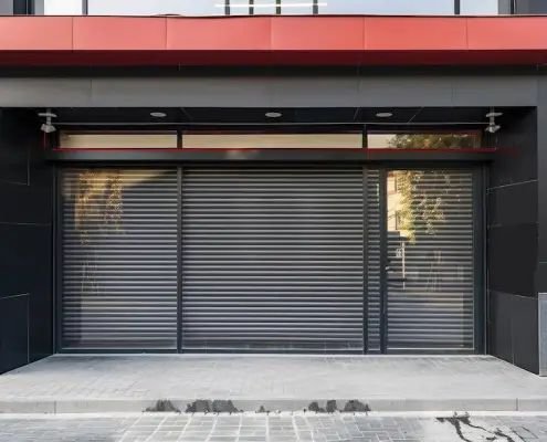 Modern storefront facade with a wide gray roller shutter door, framed by black panels and a red awning over a paved entry.