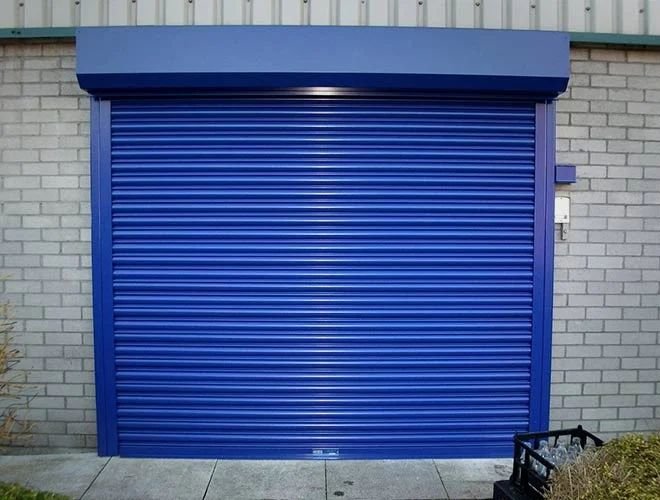 Blue metal roll-up door installed on a commercial building, featuring a ribbed shutter curtain and matching frame against a gray brick wall.