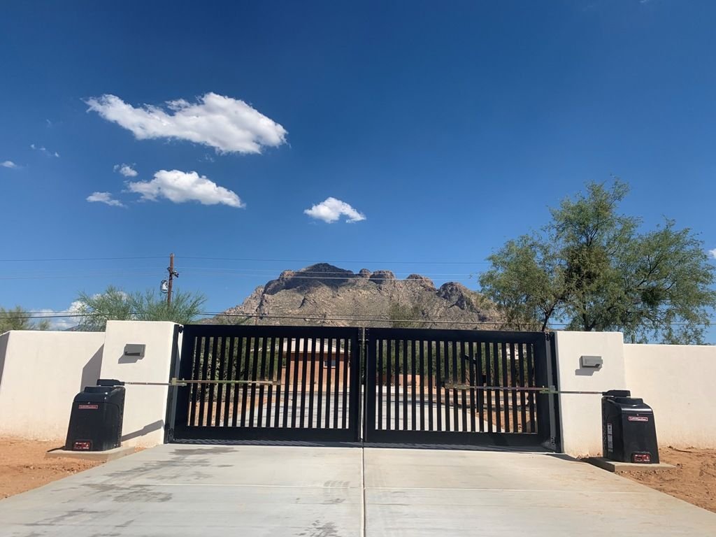 Desert property entrance with a black vertical-slat driveway gate, stucco walls, access control boxes, and mountain views under a blue sky.