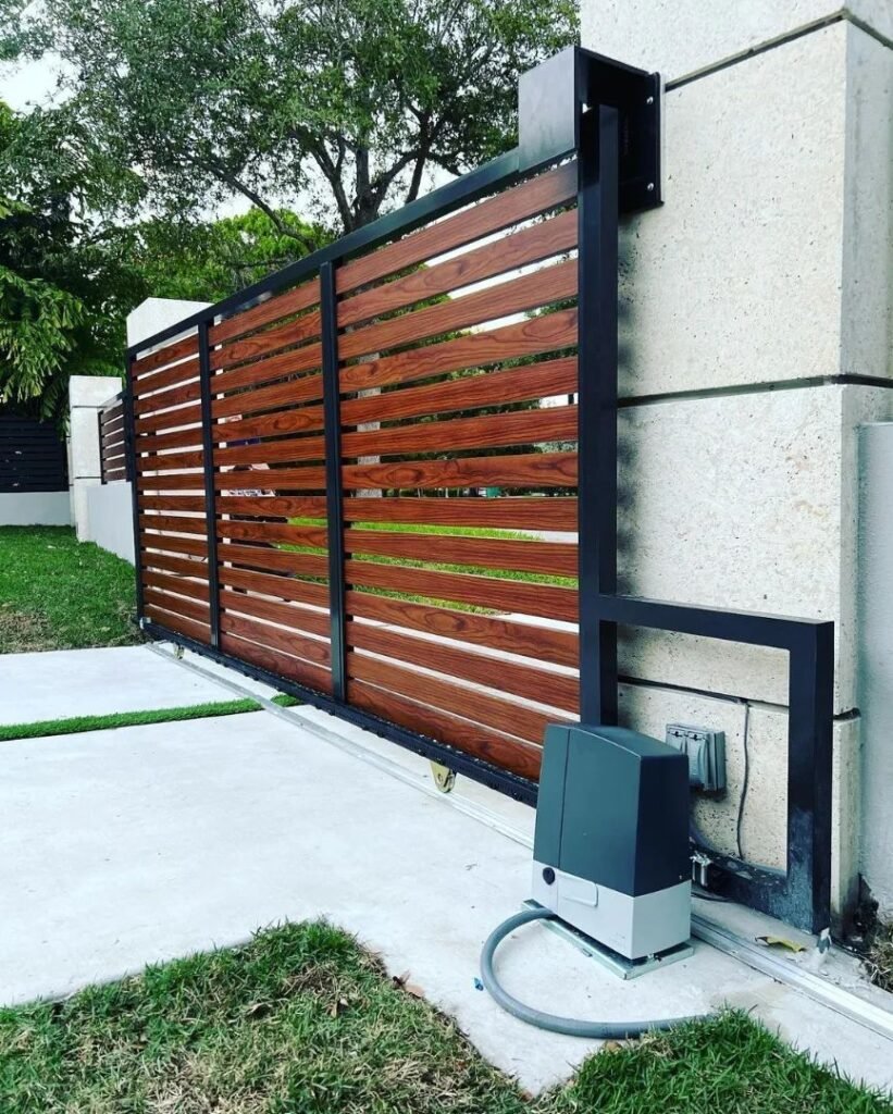 Automatic wood-and-steel sliding driveway gate with horizontal cedar slats, installed on a residential entrance with a visible gate motor and concrete drive.