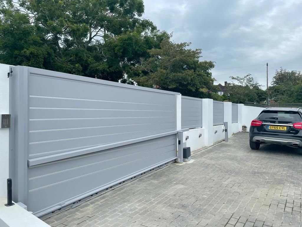 Modern gray sliding driveway gate with horizontal privacy panels and an automatic opener, installed along a long boundary wall on a paved residential driveway with a parked car.