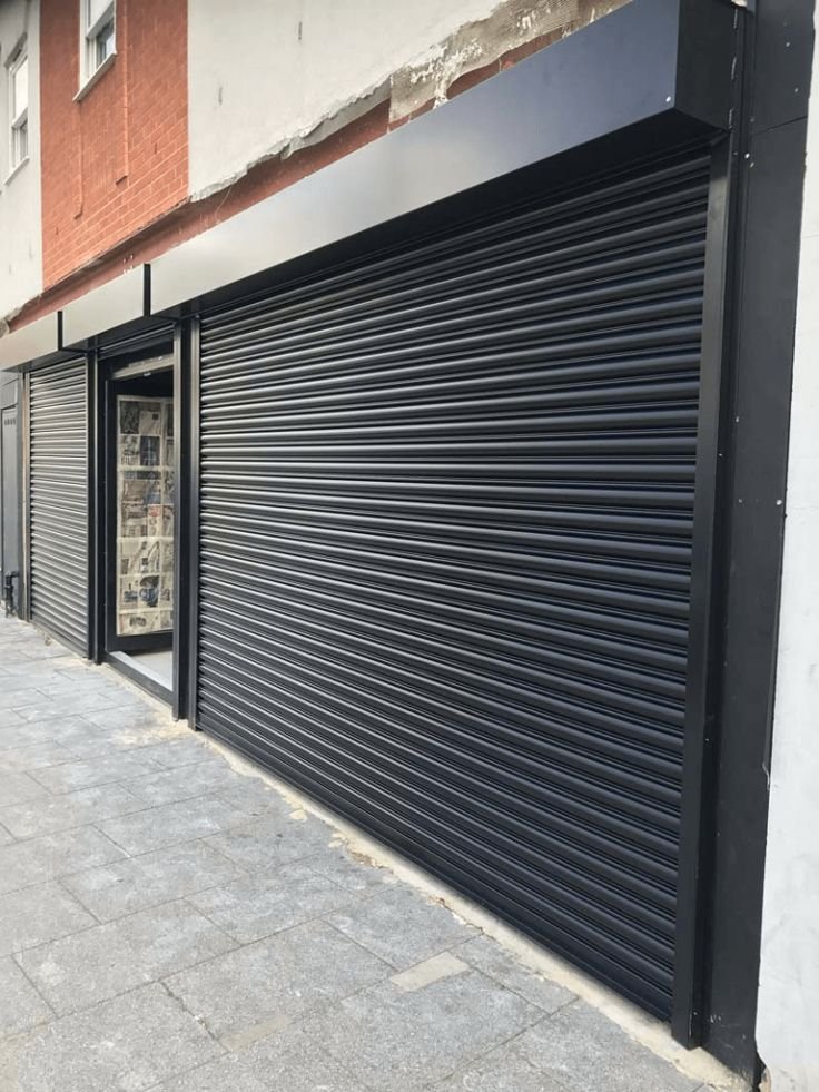 Retail storefront with a black metal roll-up door closed over a wide opening, next to a glass entry door and framed by modern black cladding.